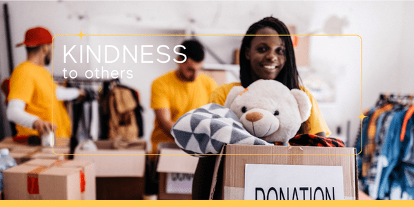 Three diverse volunteers in matching yellow shirts organizing general donations. A woman in the foreground holds a box with a teddy bear, showing how to be selfless and spread kindness through giving.